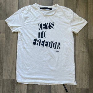 Lager 157 vit t-shirt  - Snygg vit t-shirt från The Black Label Collection med svart tryck 'KEYS TO FREEDOM' på framsidan i storlek XL. Modellen är 167 cm lång, väger 63 kg. Längden är 75 cm, axeln är 53 cm
