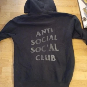 Svart hoodie Anti Social Social Club - Svart hoodie från Anti Social Social Club med stor logga tryckt på ryggen. Klassisk modell med huva och långa ärmar, perfekt för en streetwear-look. Mjuk insida och avslappnad passform.