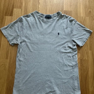  Polo Ralph Lauren T-shirt  - Snygg grå t-shirt från Polo Ralph Lauren med klassisk passform och korta ärmar. T-shirten har en broderad marinblå logga på bröstet och är tillverkad i mjuk bomull. Är även helt perfekt för sommaren nu när det börjar bli varmare och soligt. Skrev om ni har några frågor eller funderingar!