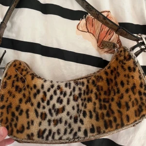 Leopardmönstrad axelväska med ormskinnsdetaljer - Cool axelväska i leopardmönstrad pälsimitation med bruna och svarta toner. Väskan har en böjd form, dragkedja upptill och detaljer i ormskinnsmönstrat syntetmaterial på botten och axelremmen. Perfekt statement piece för att lyfta din outfit.