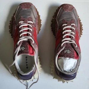 Eytys Fugu spikiga sneakers i rött och grått - Unika sneakers från Eytys med chunky, spikig sula i brunt och vitt. Ovandel i rött tyg, grått och vinrött mocka, samt lila detalj på hälen. Skorna har snörning och cool retrovibe, perfekta för dig som vill sticka ut. De är Sample så de är ganska unika och sällsynta.
