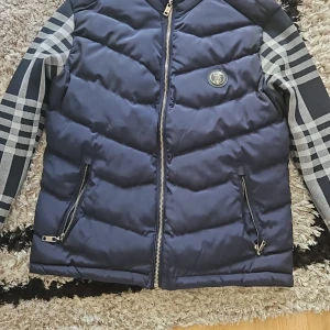 Blå pufferjacka  - Säljer en blå pufferjacka med klassiskt rutiga ärmar och insida. Jackan har dragkedja framtill, två fickor med dragkedja och ikonisk B logga på bröstet och ryggen. Perfekt för kalla dagar och riktigt snygg streetstil.