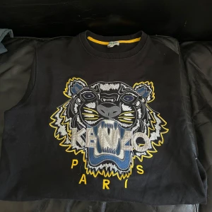 Svart Kenzo tröja med tigerbrodyr - Svart tröja från Kenzo med ikonisk tigerbrodyr framtill i grått, blått och gult med tydlig Paris-text under tigern. Perfekt för dig som vill sticka ut med en streetstyle-look.