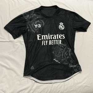 Real Madrid x Y-3 svart fotbollströja - Svart Real Madrid x Y-3 fotbollströja med vita tryck, rosdetaljer och loggor på bröstet. Tröjan har korta ärmar, rund hals och är tillverkad i ett lätt, tekniskt material (HEAT.RDY) som andas. Snygg design med subtila blommönster och ikoniska klubbmärken.