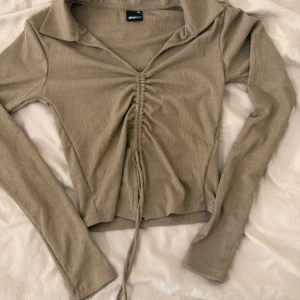 Beige ribbad långärmad topp - Trendig beige långärmad topp med ribbad struktur och snygg snörning framtill, aldrig använd. Djup v-ringning med krage och croppad passform. Perfekt för att lyfta din outfit med en cool detalj framtill.