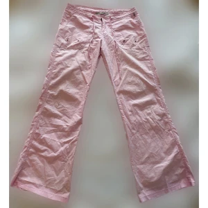 Ljusrosa utsvängda byxor med snörning - Lågmidjade tunna byxor med snörning i midjan, stora fickor fram och bak. Innerbensmått: 81cm. Midja rakt över: 42cm. Modellen på bilden är 157cm