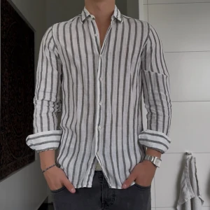 Randig linneskjorta från Massimo Dutti - Snygg vit skjorta i 100% linne från Massimo Dutti med vertikala grå ränder. Skjortan har klassisk krage, knäppning framtill och långa ärmar. Perfekt för en fräsch och stilren look under varmare dagar.