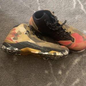 Under armour Steph curry retros  - Snygga Under Armour Curry One basketskor med högt skaft, svart och guld bas med röda detaljer och coolt randigt mönster. Sulan har kamouflageinspirerat mönster i grått, vitt och guld. Skorna har snörning och är perfekta för basket eller streetstyle. 