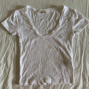 Vit ribbad topp med v-ringning - En basic vit ribbad topp från Urban Outfitters med djup v-ringning och korta ärmar. T-shirten är figurnära och har en mjuk, stretchig känsla som gör den superbekväm. Perfekt att styla med jeans eller kjol för en clean look.