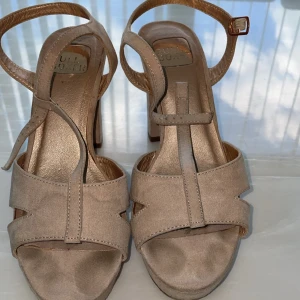 Beige sandaler med klack från Bullboxer - Snygga beige sandaler från Bullboxer med hög klack och plattform. Skorna har öppen tå, breda remmar i mocka och justerbart spänne runt ankeln. Perfekta för dig som vill ha en stilren och trendig look med extra höjd.