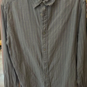 Blå randig linneskjorta med knappar - Snygg blå linneskjorta med tunna vita ränder och klassisk krage. Skjortan har långa ärmar och knäppning framtill. Perfekt för dig som gillar en stilren och enkel look.