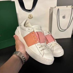 Valentino Garavani open sneakers  - Snygga Valentino Garavani Rockstud sneakers i vitt skinn med rosa och aprikosfärgade detaljer. Klassiska nitar baktill och breda band över snörningen ger en unik look. Perfekta för dig som vill sticka ut med exklusiva sneakers.