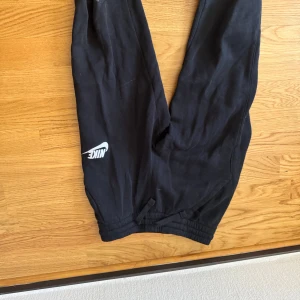 Svarta mjukisbyxor från Nike - Svarta mjukisbyxor från Nike med vit logga på benet. Byxorna har resår och snörning i midjan för skön passform. Standard fit och tillverkade i mjukt bomullsmaterial. Perfekta för chill eller träning.