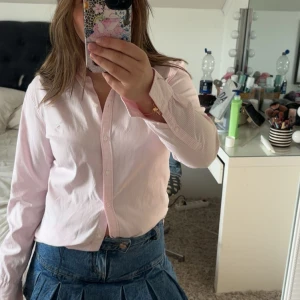 Ljusrosa randig skjorta med knappar - Säljer en ljusrosa skjorta med tunna vita ränder och klassisk krage. Skjortan har långa ärmar och knäppning framtill. Perfekt att styla med jeans eller kjol för en fräsch look. Materialet känns mjukt och lätt, troligen bomull.