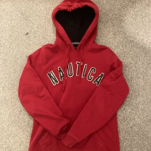 NAUTICA hoodie - En röd NAUTICA hoodie med mörkblåa bokstäver. Bokstäverna på tröjan SKA SE UT SÅ och det är själva modellen på tröjan. Har använt dem ett fåtals gånger men kommer inte längre till användning. Skriv gärna vid några funderingar eller frågor!!!!