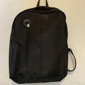 En svart enkel lite ryggsäck med flera fack i och utanför. Med band som går att göra längre eller kortare. 27 cm hög och 25 cm bred.