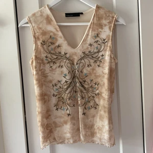 Topp - En superfin beige topp med brun tie dye effekt och bruna och blå pärlade detaljer på i ett coolt mönster. Toppen har även luttuce edge på nederkanten Toppen är från kappahl och ser ut och vara från 2000 talet