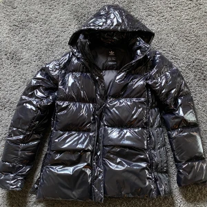 Adidas Glossy dunjacka strlk xs  - Adidas Originals Glossy A-shape down puffer jacket. Strlk XS (32). Dunjacka. Knäppning på sidorna. Två fickor fram, en innerficka. Avtagbar luva. Inköpt december 2021 och är sparsamt använd. (Kvitto finns)