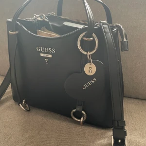 Guess väska - Jättesnygg väska från Guess som tyvärr inte används längre. Väskan är i fint skick med endast lite färgskav på ”baksidan” på metallen (syns på bilden). Fint skick på sidan i tyget! 