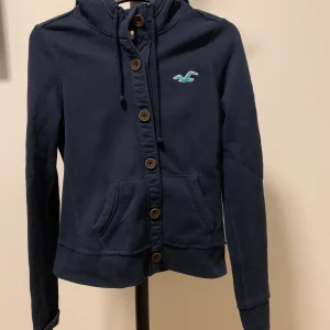 Hollister kofta  - Blå hollister kofta med luva i storlek M men passar S och XS också. Jätte fina unika knappar samt tryck på ärmen och bröstet. I bra skick. 