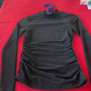 Svart glittrig långärmad top - Materialet är 59%polyamid,31%metallisk fiber, 10%elastan Längd 59 cm som längst Rynkade sidor , lång ärm o liten polokrage  Använd ett par gånger Bra skick  