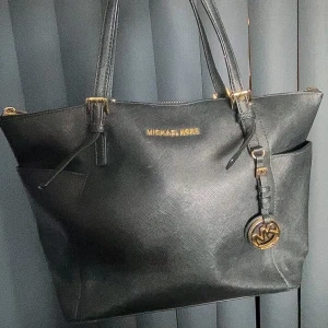 Michael Kors, väska  - Jet Set Item Shoppingväska korniga nötkött läder svart Produktbeskrivning två fickor på sidorna fötter av metall stängs med blixtlås två handtag (henkel fall 21 cm) polerad guldfärgad metall avtagbar hänge Material: korniga nötkött läder Vikt: 0,7                                                                                 Haft i ca 2 år men är knappt använd                               Ordinarie pris : 2 700kr                                                       Priset är diskuterbart 