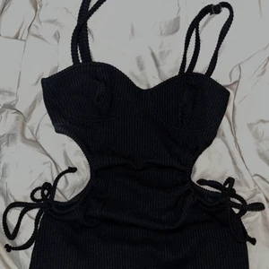 Klänning NA-KD - Svart klänning från NA-KD med öppning vid sidan av revbenen som man kan justera själv med band som går att knyta om hur man vill. Klänningen går ner till knäna. Storlek XXS men passar XS/S. Oanvänd. 