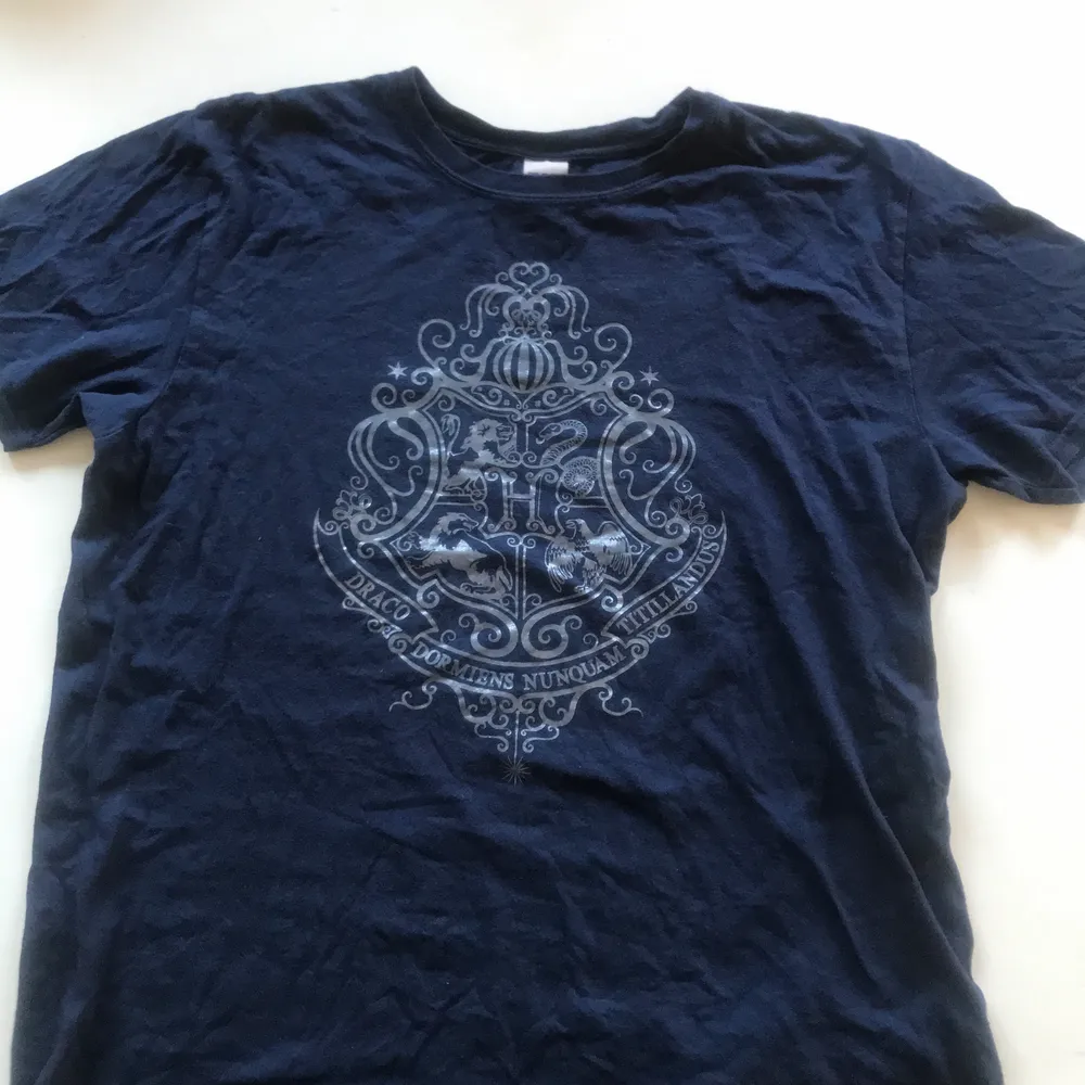 Mörkblå t-shirt med silvrigt hogwarts tryck. Köpt på sci-fi bokhandeln.  T-shirten är i begagnat skick men inte väldigt använd. Den är gjord av 100% bomull och är i storlek L.. T-shirts.