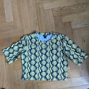 T-shirt  - T-shirt i blankt stretchigt material. Gul, ormskinnsmönstrad. Hämtas upp på södermalm 