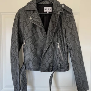 Skinnjacka - Fake skinnjacka i storlek XS från NAKD i grått/svart ormskinnsmönster. Skärpet går att ta bort och jackan går att stänga om man skulle vilja det :) Kan frakta (även spårbart) eller mötas upp i Göteborg 🤗