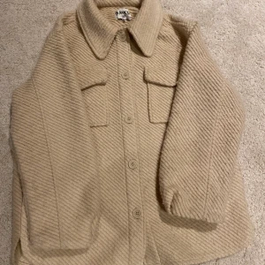 OverShirt i Ull - En beige overshirt i ullblanding i en längre modell. Overshirten har ett randigt mönster och två fickor på bystet, samt en gömd ficka på vardera sida. 