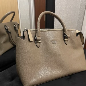 Guess väska beige  - Guess handväska beige. Sparsamt använd men dessvärre fått några skrapmärken på hörnen samt handtagen. Detta är dock ingenting som är märkbart synligt.   Längd: 30 Höjd: 20 Bredd: 13