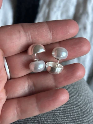 Silvriga örhängen med pärlor - örhängen i silverfärg med halvrunda former och vita pärlor. Designen är modern och stilren med en cool kontrast mellan metall och pärla. Perfekta för dig som gillar att sticka ut med accessoarerna. Märke ej angivet.