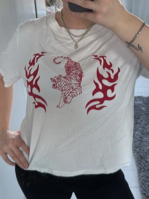 Vit t-shirt med röd tigerprint - Säljer en vit t-shirt med rött tryck av en tiger och tribal flames på bröstet. T-shirten har korta ärmar och en avslappnad passform. Perfekt för dig som gillar streetstyle och vill sticka ut lite extra.