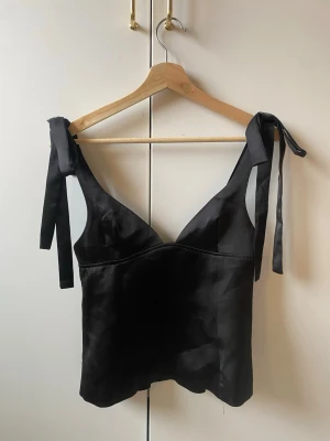 Svart satinlinnetopp med knytband - Stilren svart linnetopp i satin med breda axelband som knyts på axlarna. Toppen har en djup v-ringad urringning och en figurnära passform. Perfekt att styla med jeans eller kjol för en trendig look. aldrig använd!