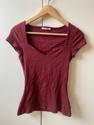 Vinröd ribbad topp med v-ringning - vinröd ribbad topp med snygg v-ringning och korta ärmar. Toppen har en figurnära passform och är tillverkad i mjukt material. Perfekt att styla med jeans eller kjol för en trendig look. gott skick!!