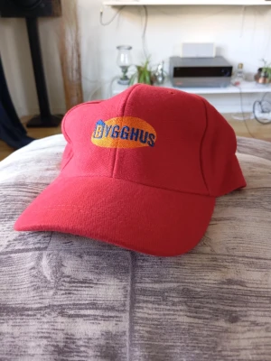 Ny Röd keps med BYGGHUS-logga - Säljer en röd keps med böjd skärm och broderad BYGGHUS-logga i blått och orange framtill. Klassisk modell med justerbar rem bak. Perfekt för dig som vill sticka ut med en färgstark accessoar.