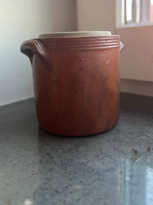 Brun keramikburk med handtag - Rustik brun keramikburk med två små handtag på sidorna och bred öppning. Ytan är matt och burken har en naturlig, jordnära färg. Perfekt för förvaring av småsaker eller som dekoration i hemmet.
