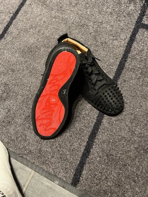 Svarta Louboutin sneakers med nitar - Säljer ett par svarta sneakers från Christian Louboutin med klassisk röd sula och svarta nitar på tån. Skorna har en låg silhuett, breda svarta skosnören och är tillverkade i mocka och läder. Perfekt för dig som vill sticka ut med en edgy och lyxig stil.