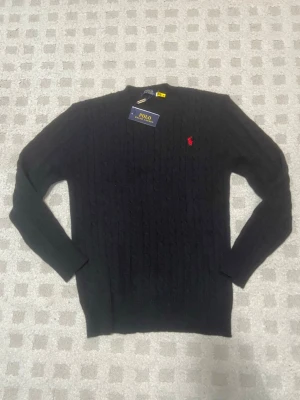 Svart kabelstickad Ralph Lauren tröja - Kabelstickad svart tröja från Polo Ralph Lauren med klassisk röd broderad logga på bröstet. Tröjan har rund halsringning och långa ärmar. Tillverkad i mjuk ull som känns riktigt skön mot huden. Perfekt för dig som vill ha en stilren och tidlös look. QR-koden samt serienumret funkar!