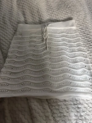 Vit stickad kortkjol med snörning - Supersöt vit stickad kortkjol med vågigt mönster och snörning i midjan. Kjolen har en mjuk och stretchig känsla, perfekt för en chill och trendig look. Passar perfekt till sommaren och stranden eller våren och är lätt att matcha med olika toppar.