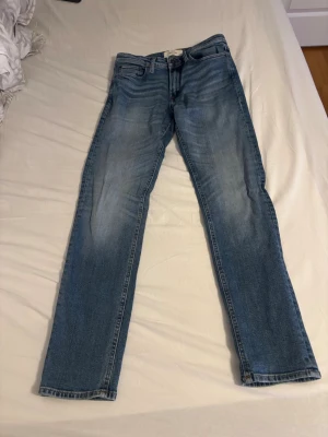Blå raka jeans med slitningar - Säljer ett par klassiska blå jeans med raka ben och lätt slitna detaljer framtill. Jeansen har fem fickor och normal midja. Perfekta till en avslappnad stil och passar till det mesta. Materialet är denim i bomull.