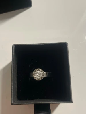 Stålring  - Stilren silverfärgad ring från Efva Attling med rund dekoration framtill, täckt av gnistrande små stenar och texten 'Peace of Sweden' runt om. Ringen har en modern och elegant design som verkligen sticker ut.