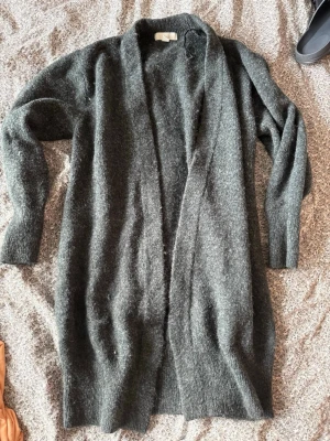 Mörkgrå lång kofta från H&M - Mysig lång kofta i mörkgrått från H&M. Koftan har öppen front utan knappar, långa ärmar och en avslappnad passform. Perfekt att slänga över en t-shirt eller topp för extra värme. Materialet är mjukt och lite luddigt, vilket ger en skön känsla.