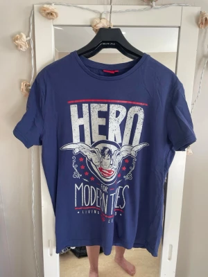 Blå Superman t-shirt med tryck - Snygg blå t-shirt med Superman-tryck och texten 'Hero for Modern Times' framtill. Klassisk rund hals och korta ärmar. Perfekt för dig som gillar superhjältar och vill ha en avslappnad stil. Tillverkad i mjuk bomull för skön känsla.