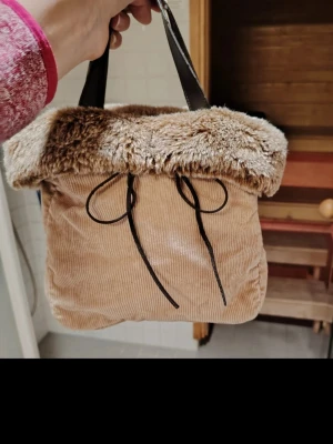 Beige handväska med fuskpälsdetalj - Söt beige handväska i manchestertyg med mjuk fuskpäls upptill och svarta handtag. Väskan har dekorativa svarta band framtill och en mysig vinterkänsla❄️. Perfekt accessoar för att lyfta din outfit❤️