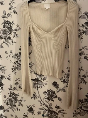 Ribbad beige topp med lång ärm - Trendig ribbad topp i beige med lång ärm och markerad byst. Toppen har en djup, fyrkantig urringning och är tillverkad i stretchigt, mjukt material. Snygg passform som framhäver figuren och ger en modern look.