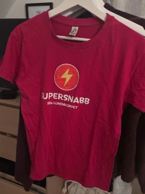 Rosa SUPERSNABB t-shirt Lilla Lundaloppet - Rosa t-shirt med tryck 'SUPERSNABB lilla LUNDALOPPET' och en gul blixt på bröstet. Klassisk rund hals och korta ärmar. Perfekt för dig som gillar sportiga och färgstarka plagg.
