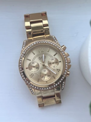 Guldig klocka med strassdetaljer - Snygg guldfärgad klocka från O.N.E med strass runt urtavlan. Superfin accessoar som funkar till allt! Kan behöva ett nytt batteri. Medföljer extra länkar.