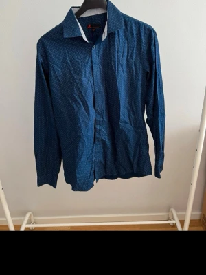 Blå skjorta med småprickigt mönster - Stilren blå skjorta med små vita prickar och klassisk krage. Skjortan har långa ärmar och knäppning framtill. Perfekt för dig som gillar en enkel men ändå snygg look. Materialet känns som bomull och ger en bekväm känsla hela dagen.